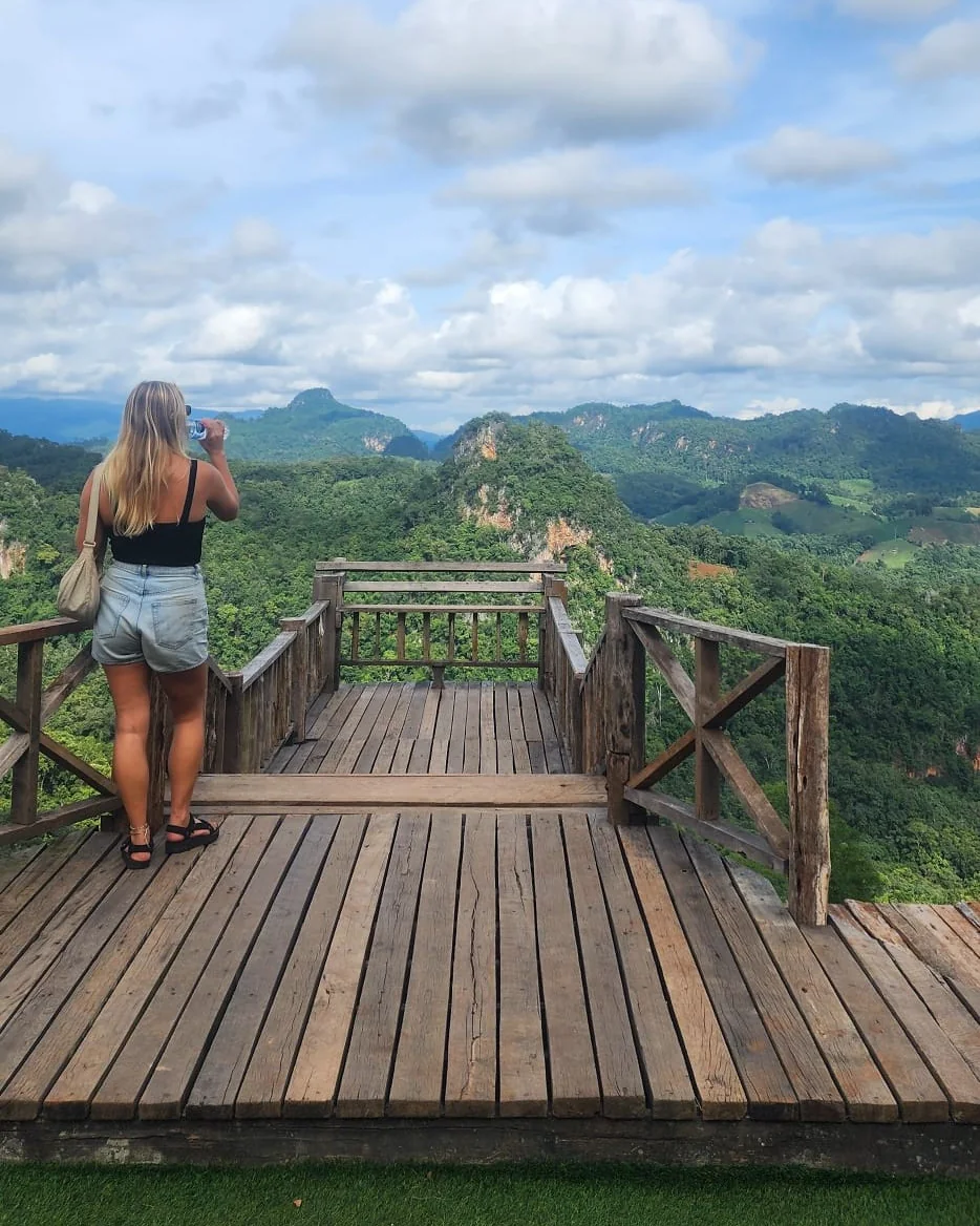 A woman with a water bottle on a wooden viewing deck, looking out over forested limestone peaks under a big sky