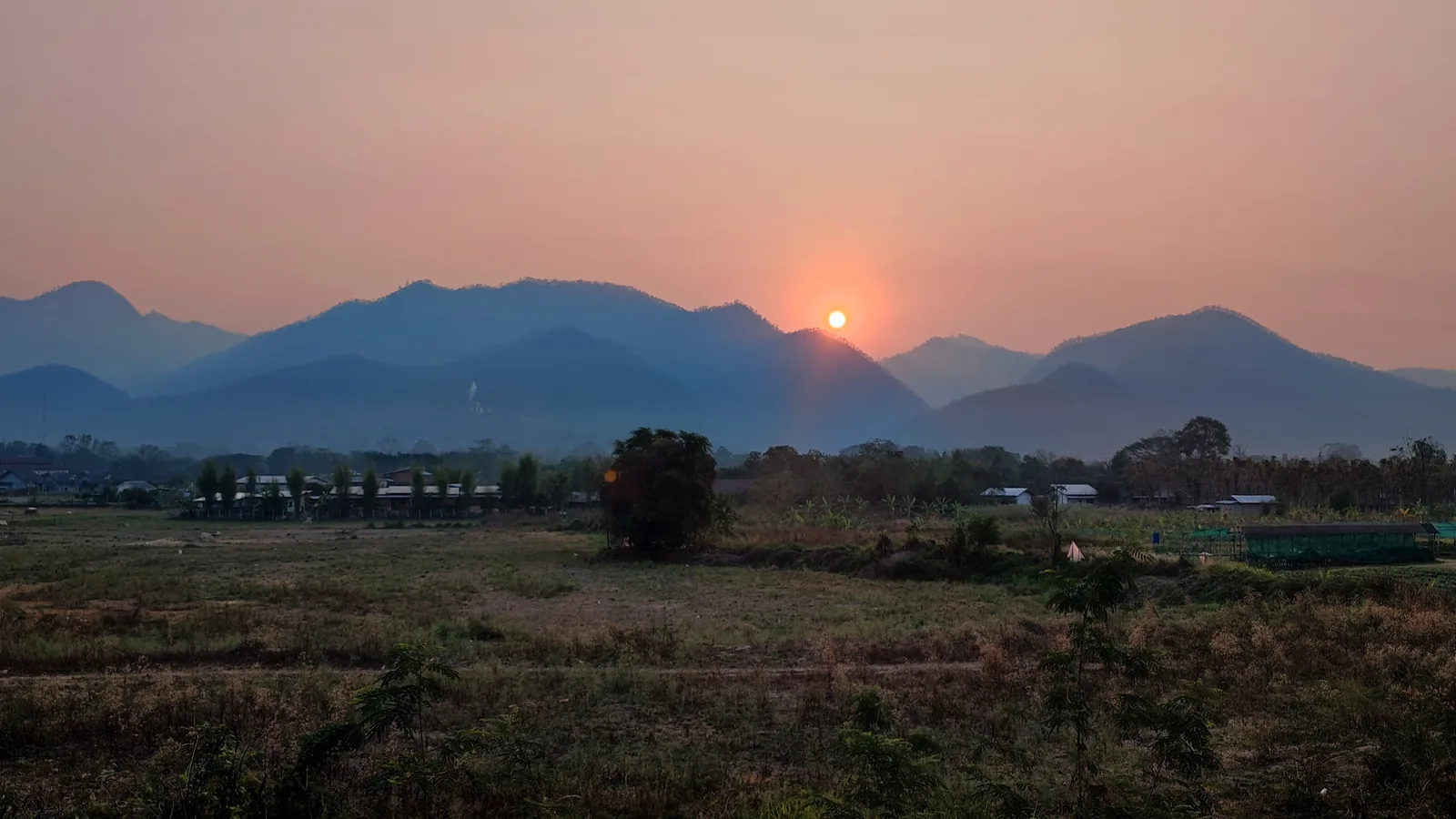 The sun rising as a small orange disc through thick pink haze over the mountains east of Pai in March, above dry fields