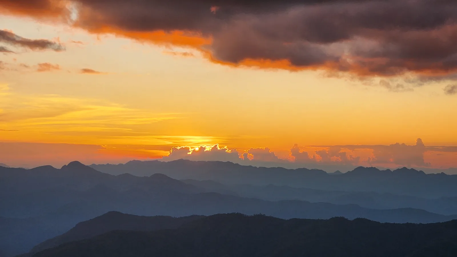 The sun setting into a bank of cloud over layer after layer of blue mountain ridges, seen from Doi Miang in December