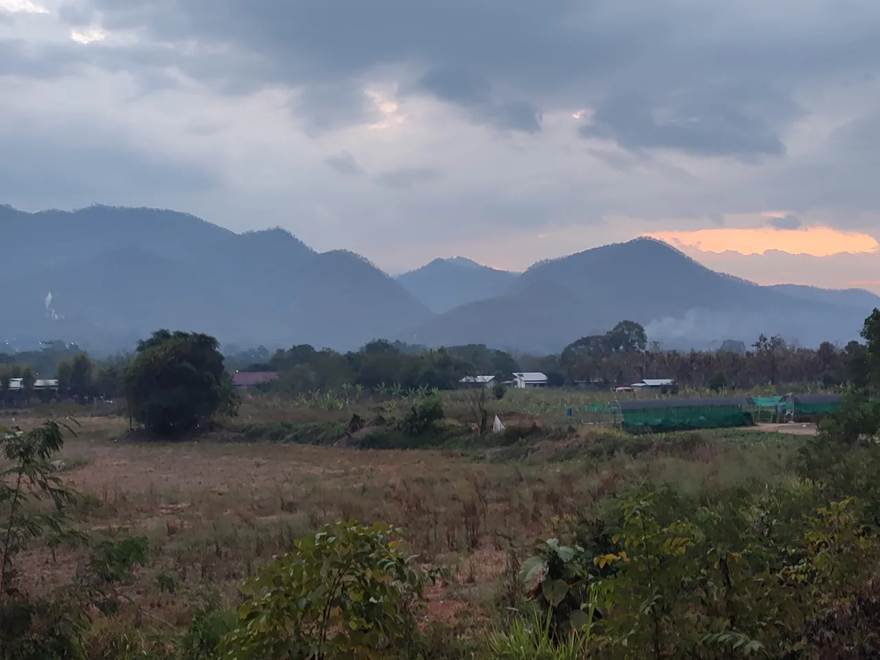 Dry brown fields on the edge of Pai at first light in February, grey cloud over the mountains and a column of smoke rising from the foot of one hill