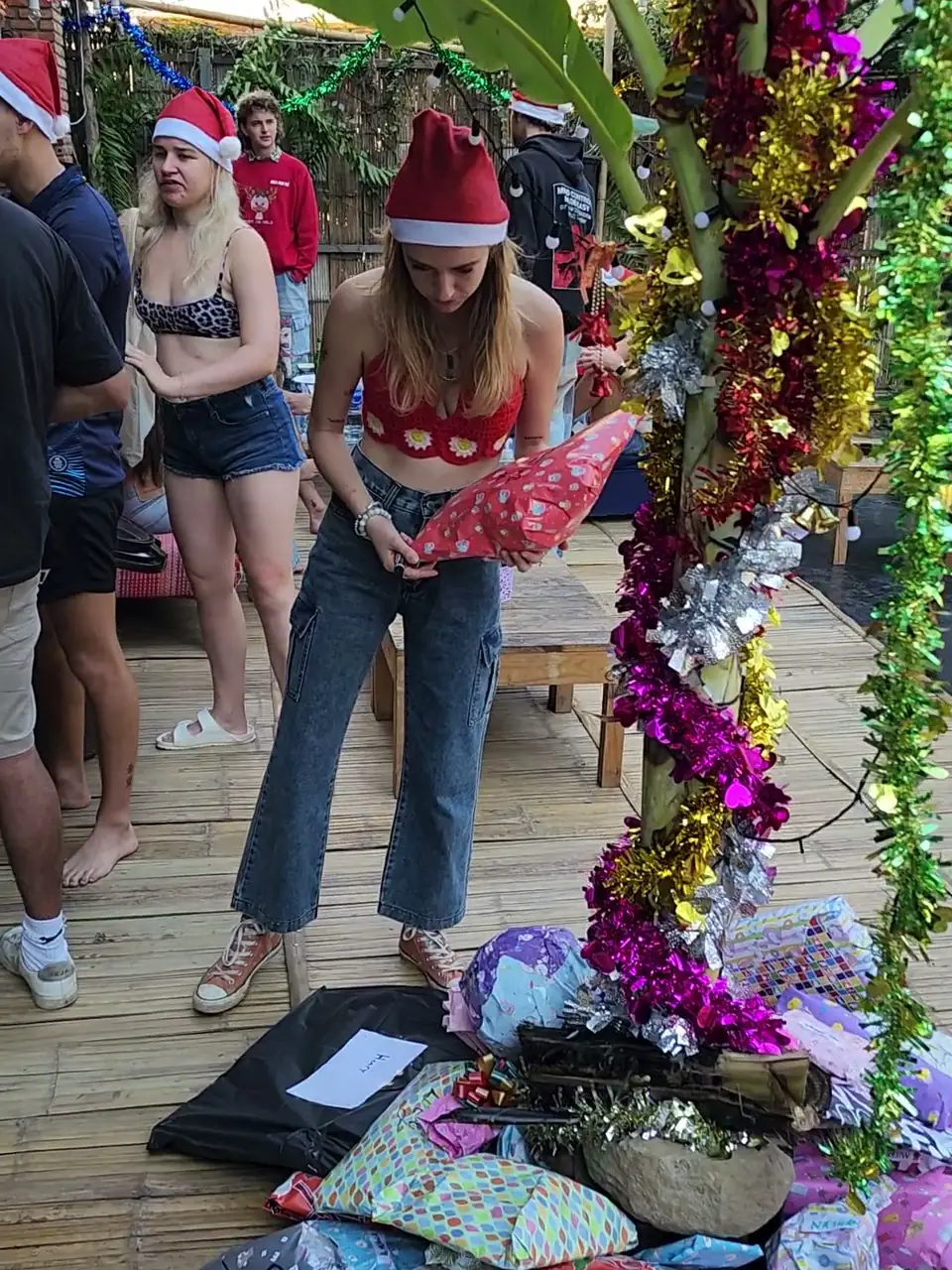 A guest in a Santa hat picking up her Secret Santa present beside the Banana Christmas Tree at Nolo Hub Pai, a banana plant wound with tinsel, with presents piled at its foot