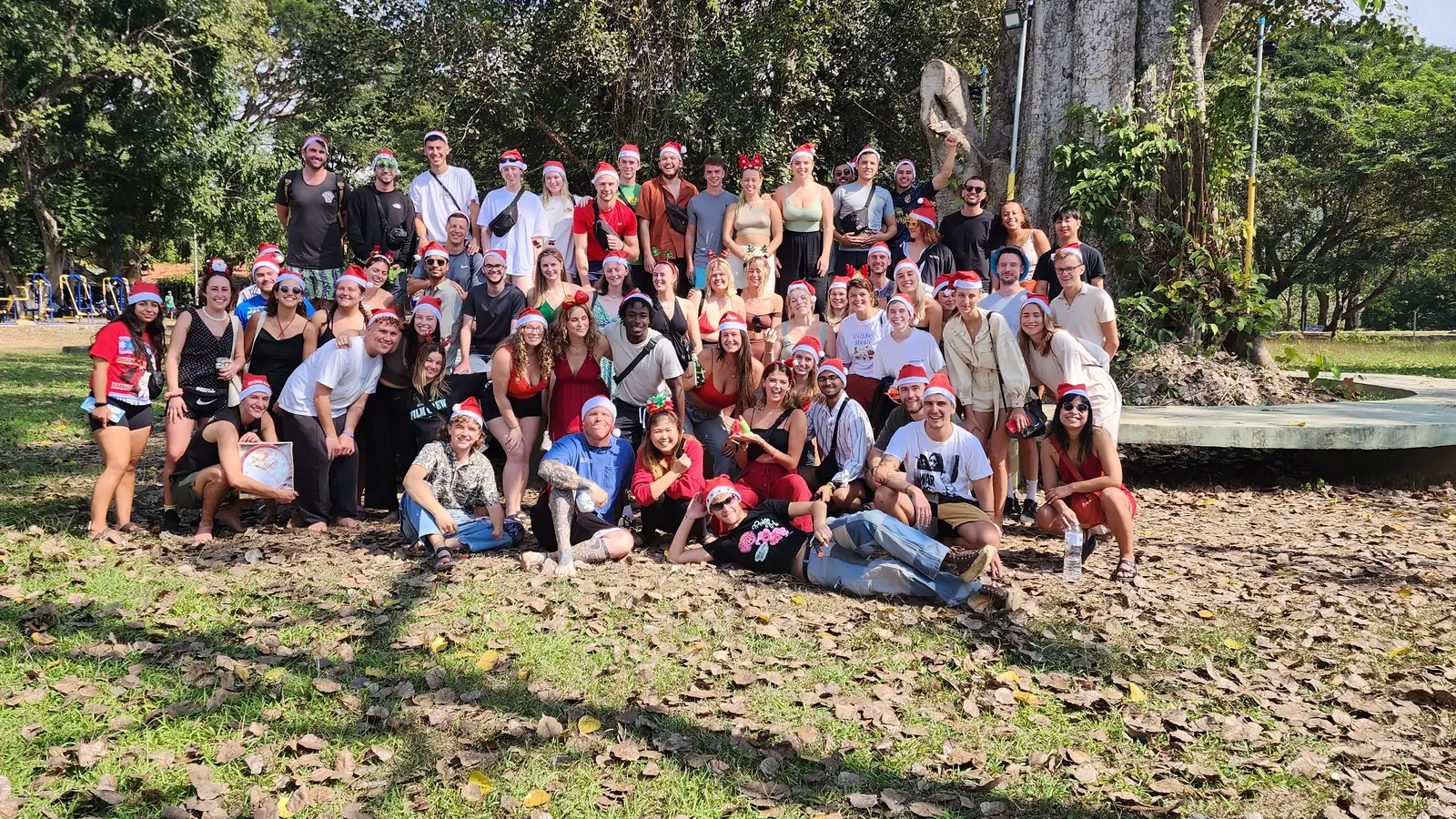 A large group in Santa hats gathered on the grass under tall trees on Christmas Day at Nolo Hub Pai