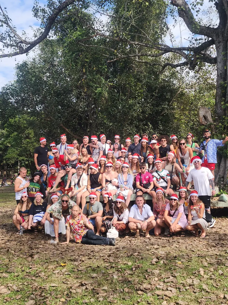 A big group in Santa hats posing on the grass under a tall tree on Christmas Day at Nolo Hub Pai