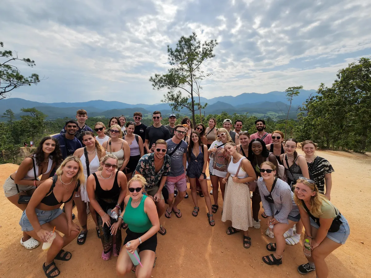 A big group from Nolo Hub Pai at Pai Canyon, crouching and smiling on the red earth, with pine trees and the mountains behind