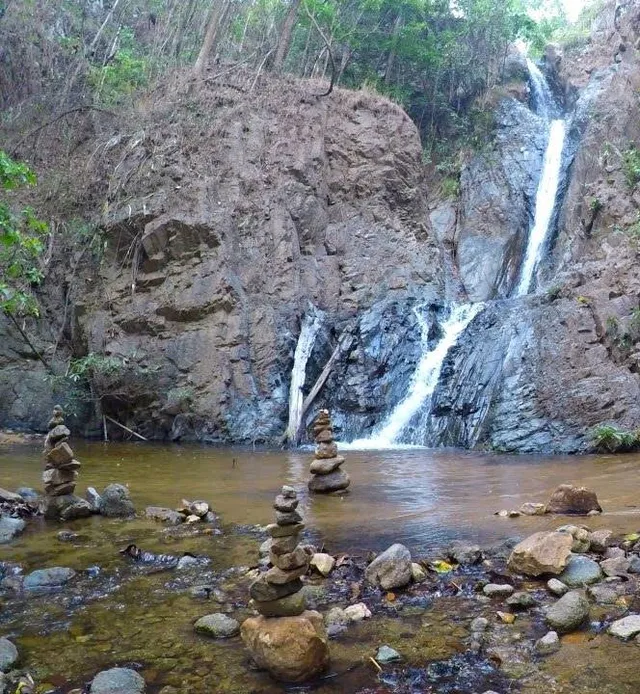 Mae Yen Waterfall falling in two streams into a shallow pool, with stacks of stones at the water's edge