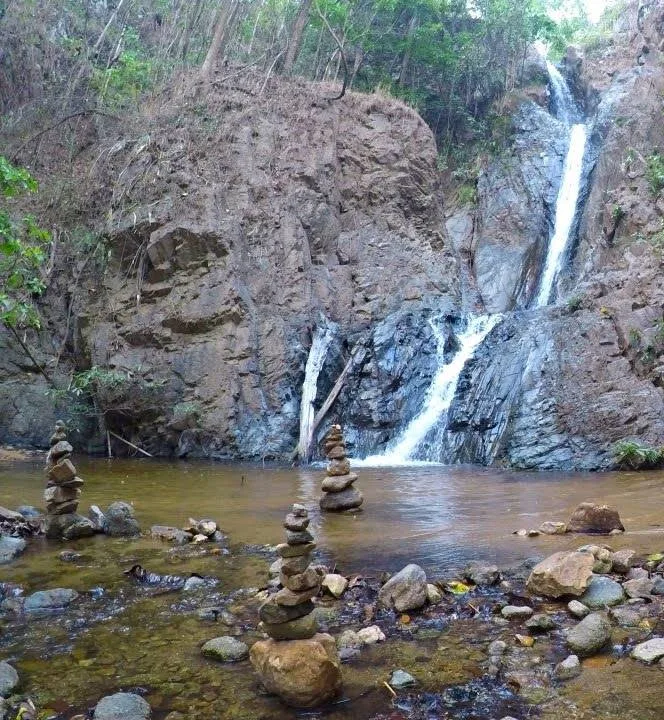 Mae Yen Waterfall falling in two streams into a shallow pool, with stacks of stones at the water's edge