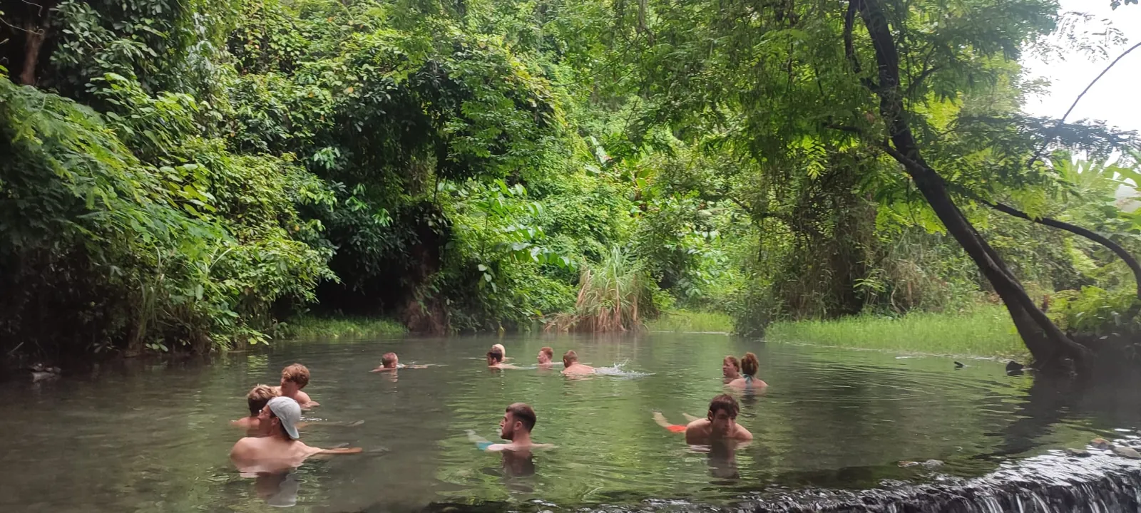 A Nolo Hub Pai group soaking in Sai Ngam's warm stream pool, with the forest behind