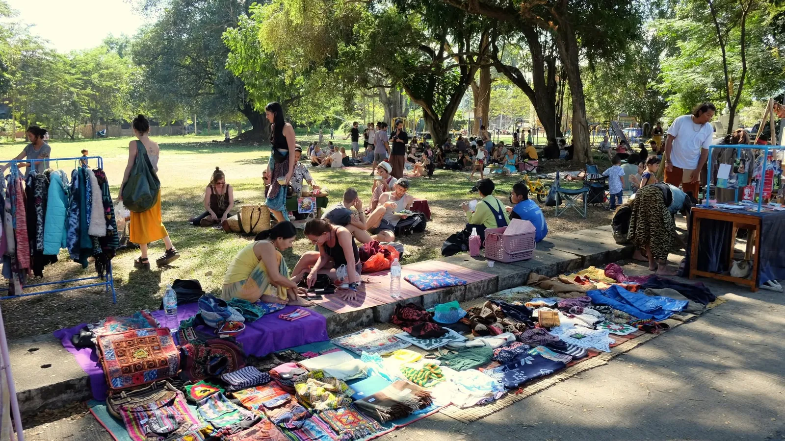 Bags, scarves and clothes laid out on mats along the path by Bodhi Tree Park, with people on the grass under the trees behind