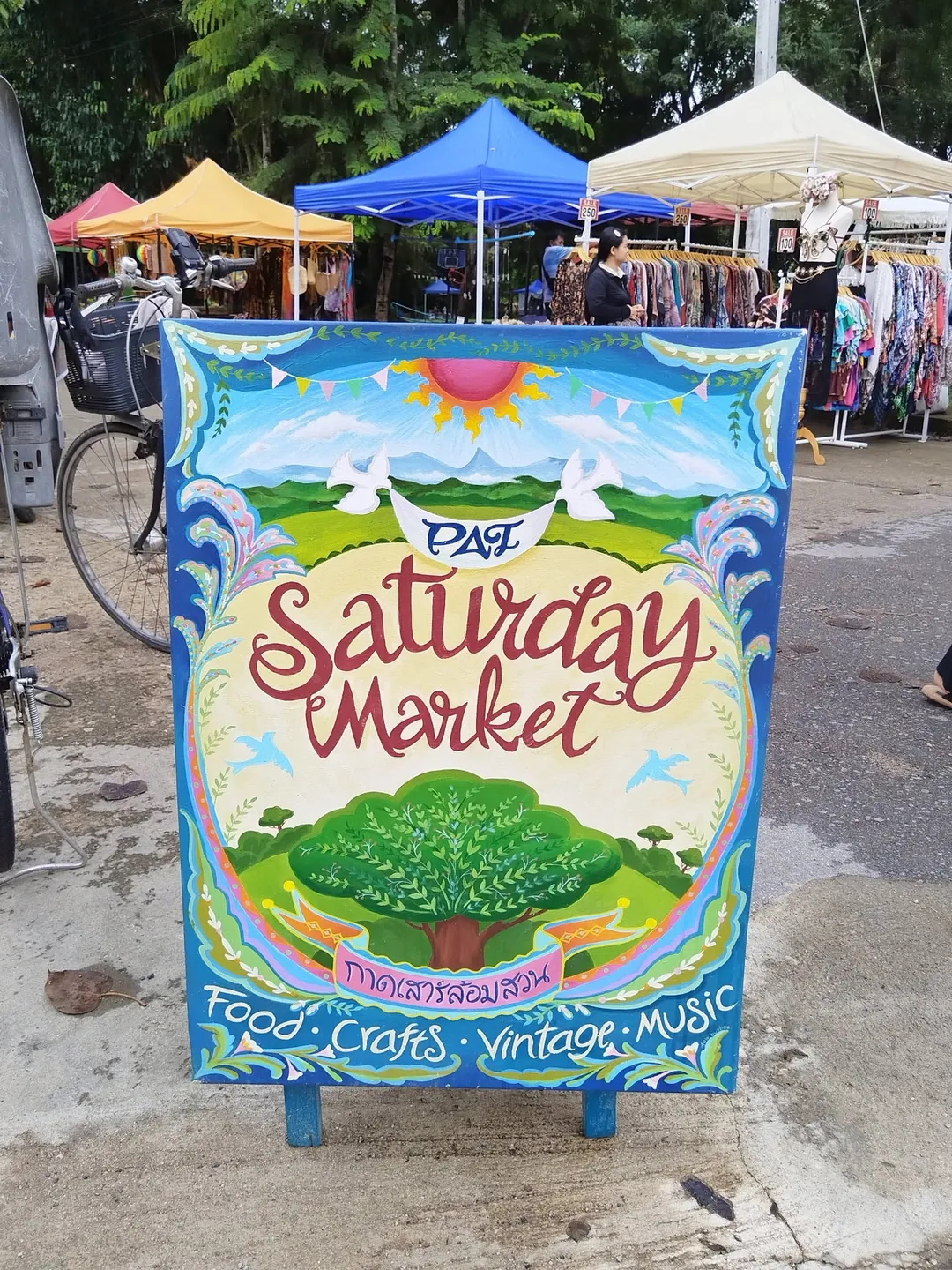 The hand-painted Pai Saturday Market sign, a big tree under a rising sun and the words Food, Crafts, Vintage, Music, with stall tents behind it