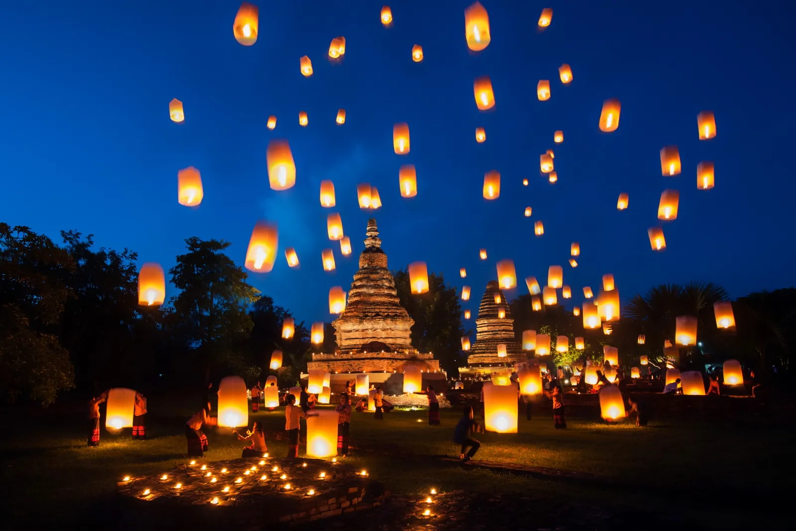 Dozens of sky lanterns rising at dusk over old brick stupas, with small lamps set out on the grass