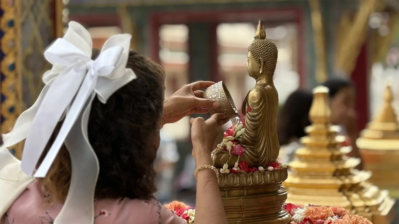 A woman seen from behind pouring water from a silver bowl over a small golden Buddha image hung with jasmine, orange carnations around its base