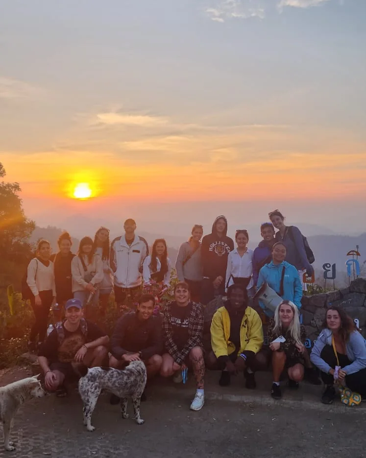 A Nolo Hub Pai group and two dogs at the Doi Kiew Lom viewpoint, with the sun going down over the mountains behind them