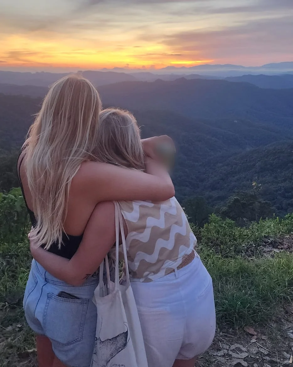 Two women hugging as they watch the sun set over layers of forested mountains