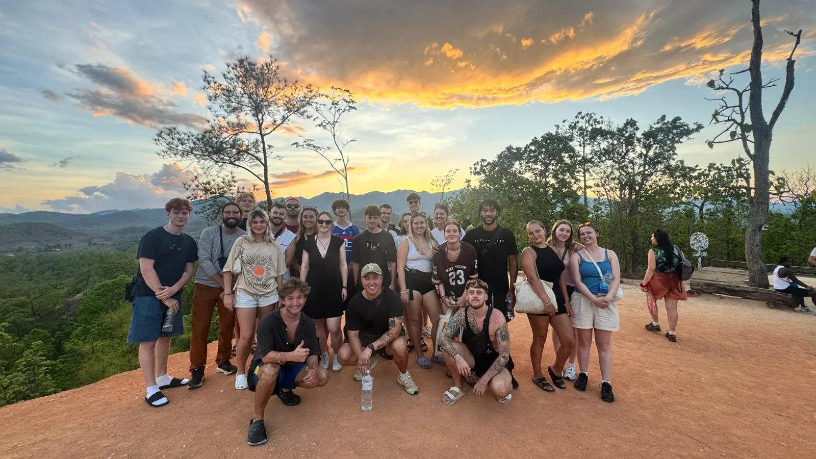 A big Nolo Hub Pai group on the red earth at Pai Canyon, under orange sunset clouds, with the forest and mountains behind