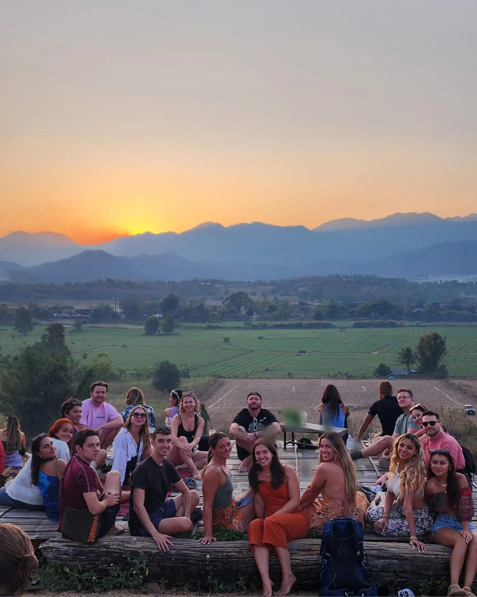 A Nolo Hub Pai group sitting on the wooden deck at Two Huts, with green fields and the sun setting behind the mountains