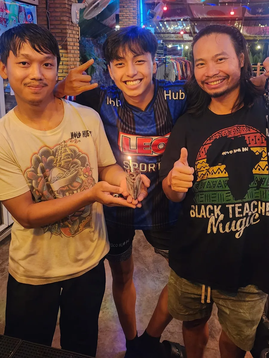 Ya, Wan and Korn together at the bar under coloured lights, two of them giving a thumbs up