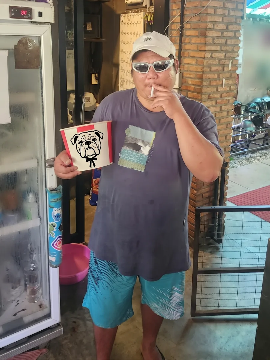 Sunny in a white cap and sunglasses by the drinks fridge, holding a bucket with a bulldog drawn on it