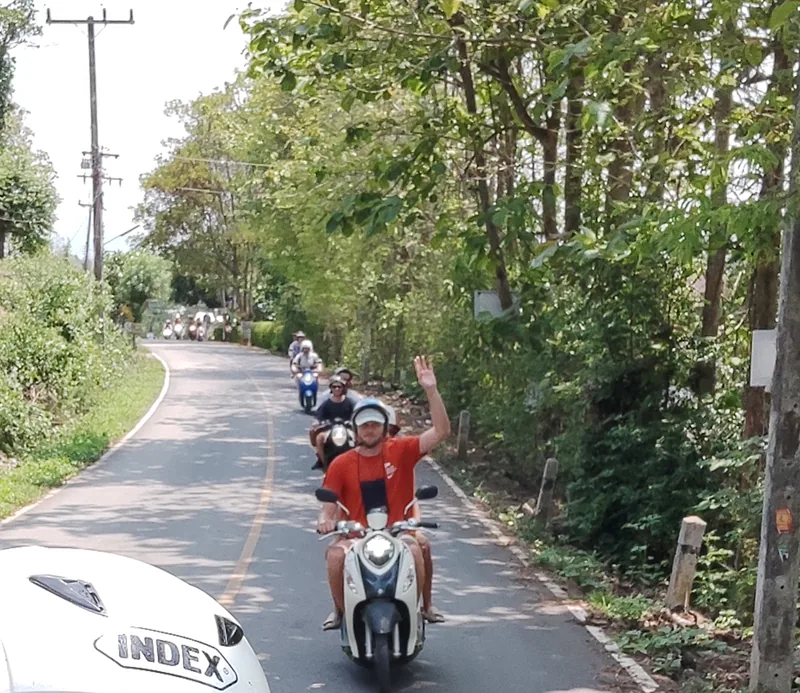 Adventure: Nolo Hub Pai guests riding scooters along a tree-lined road, the nearest one waving