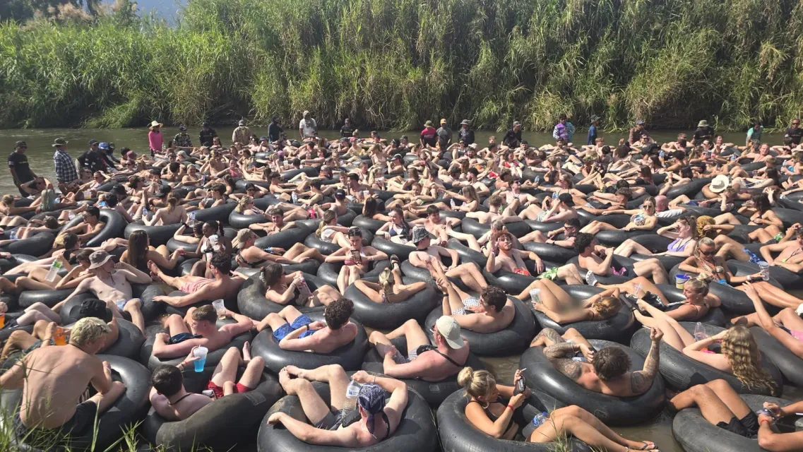 A packed crowd lying back in black inner tubes on the Pai River, guides standing in the water behind them by the tall riverbank grass