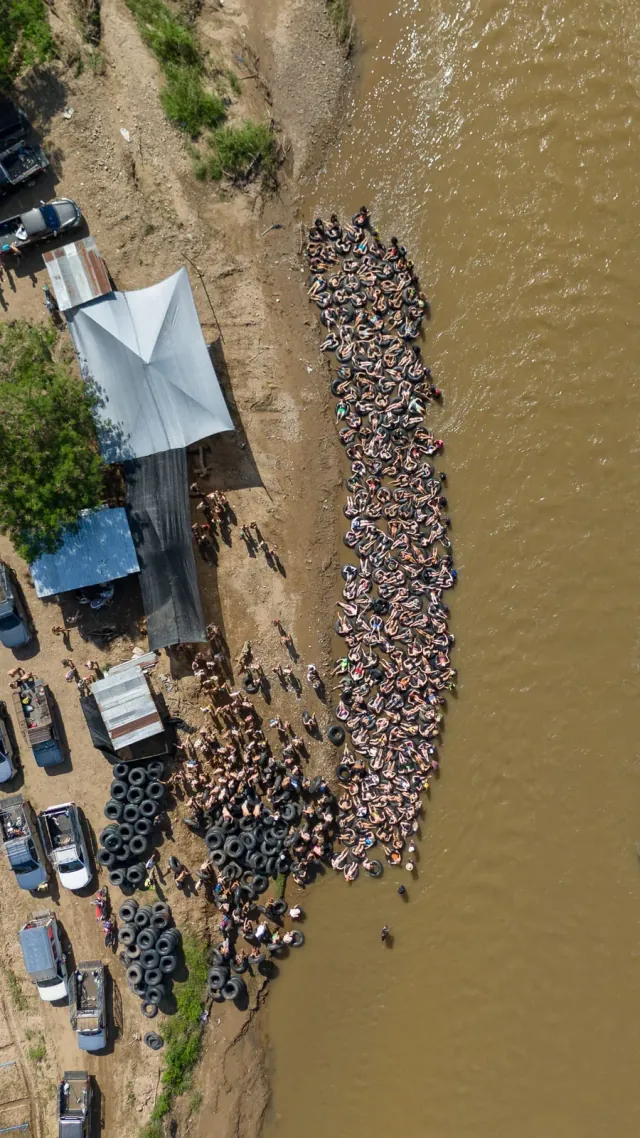 Hundreds of people in black inner tubes lined up along the Pai River's bank at the start of Tipsy Tubing, the pickup trucks parked behind, seen from straight above