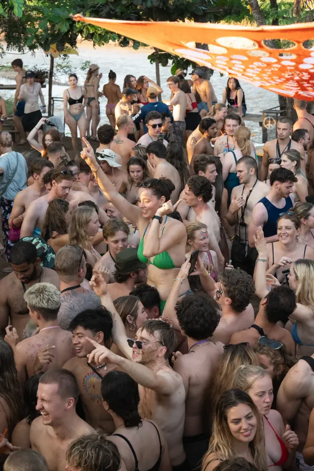 A crowd of tubers in swimwear dancing at a riverside stop under an orange shade cloth, a woman in a green bikini with her arm in the air