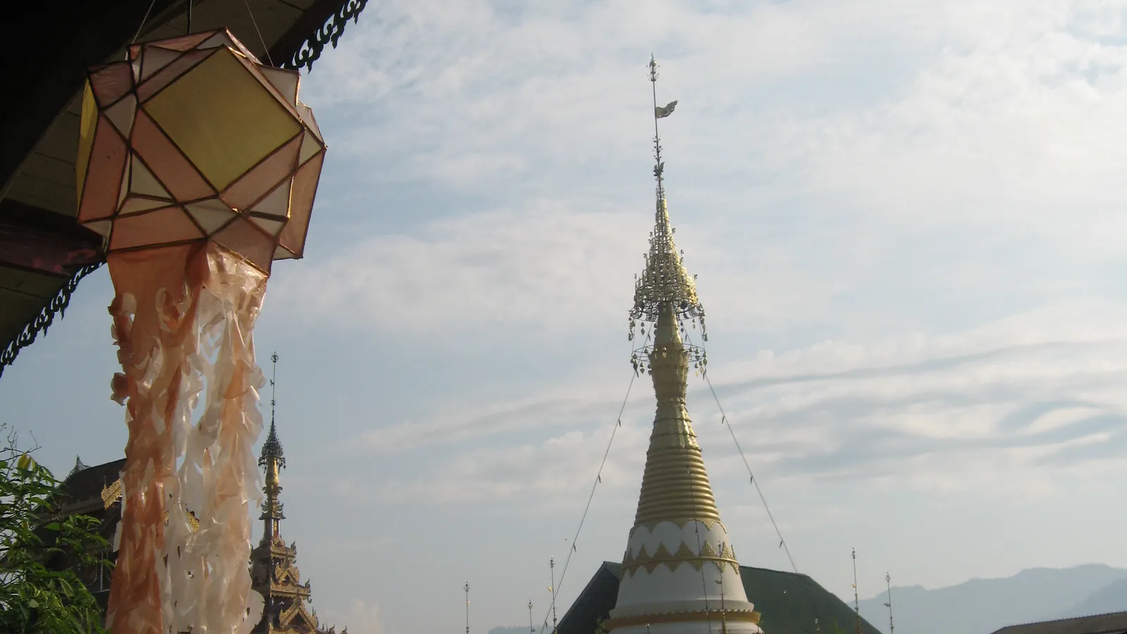 A paper lantern with long tassels hanging under a temple roof, a white stupa behind it